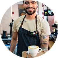 Barista hombre tatuado sirviendo café en taza blanca con máquina al fondo